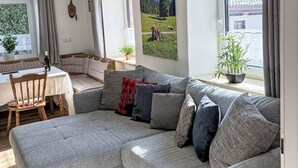 Sala de estar com sofá, mesa de jantar e cadeiras. Possui duas janelas com cortinas, um quadro na parede e um vaso com planta.
