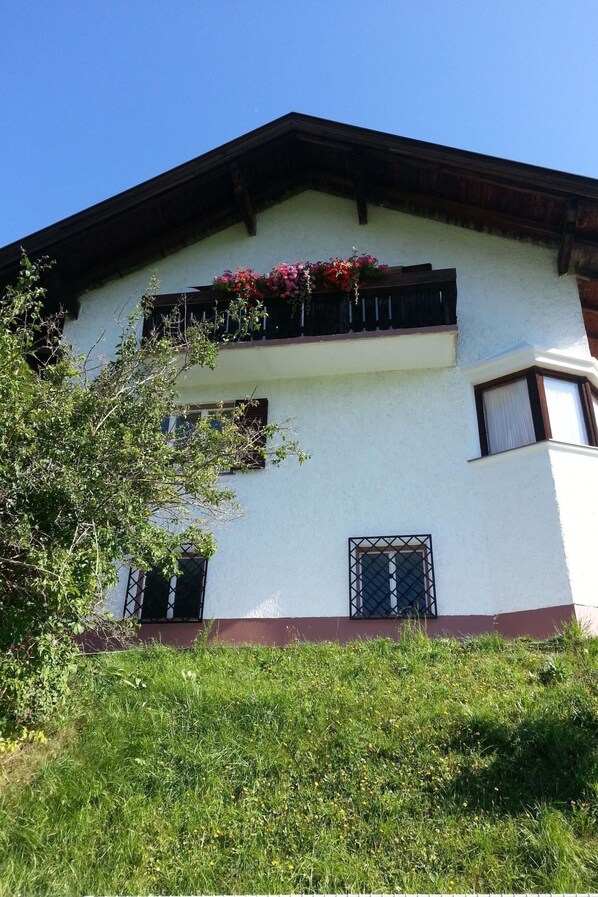 Un bâtiment à deux étages, doté d’une façade blanche, d’un toit sombre et d’un balcon orné de fleurs rouges. Il y a deux fenêtres avec des barreaux en métal et une petite porte.