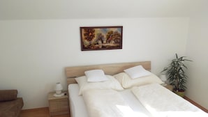 Un dormitorio con cama, mesita de noche, lámpara, un cuadro y una planta en maceta.