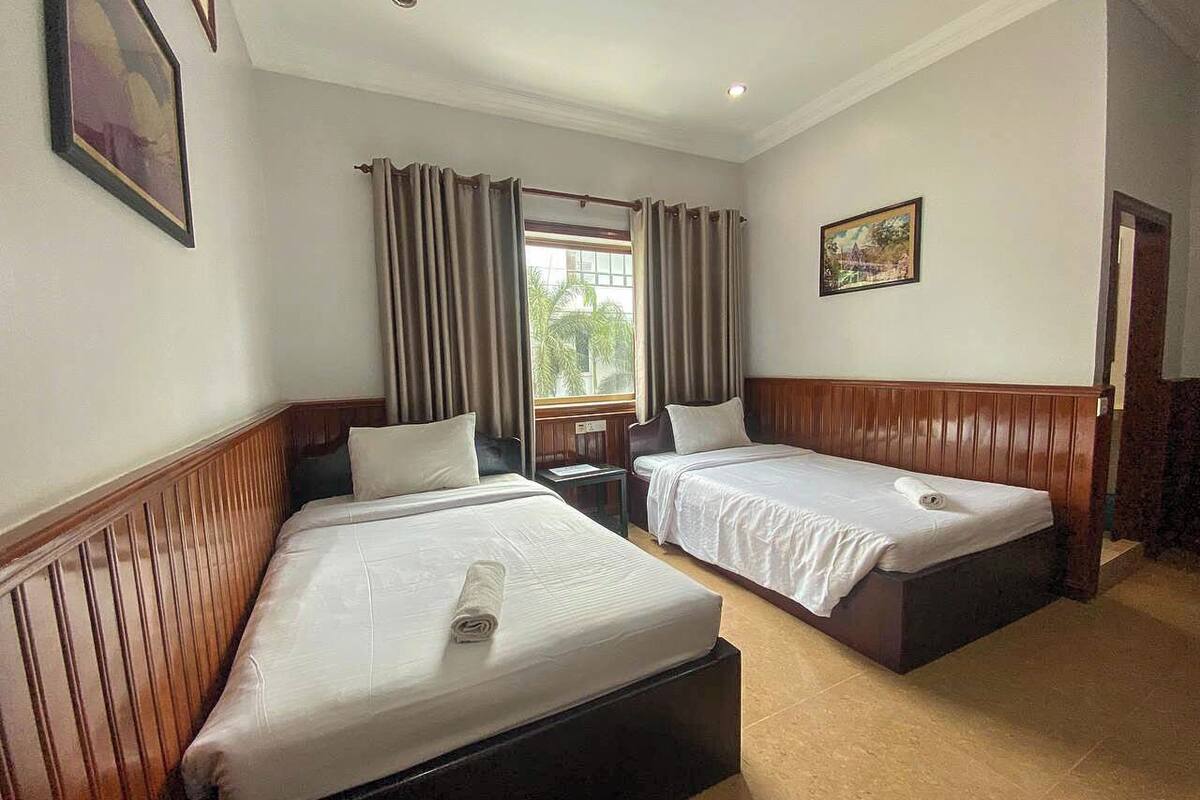A hotel room with two beds, a wooden headboard, a small table, and a window with curtains.