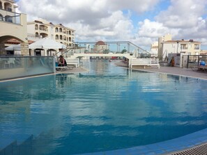 A swimming pool with lounge chairs and umbrellas, surrounded by buildings with balconies. - Modernes Ferienhaus mit Pool in Tremithousa by Interhome (Mesoyi)