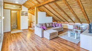 A spacious loft with a high wooden ceiling, a grey sectional sofa, and a glass coffee table.