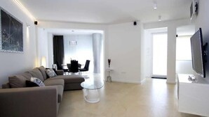 Ein modernes Wohnzimmer mit einer Couch, einem Glastisch und einem Flachbildfernseher. Im Hintergrund ist eine Essecke mit Tisch und Stühlen zu sehen.
