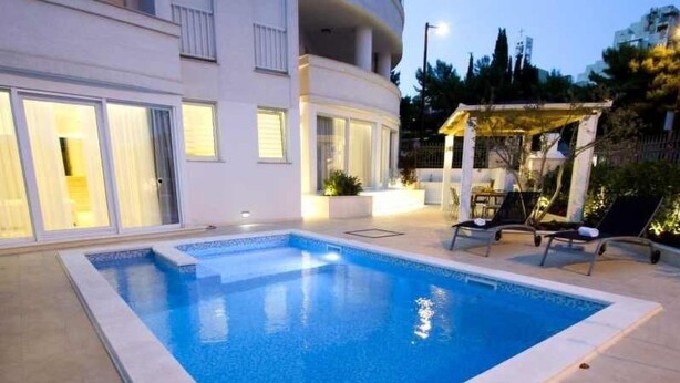Ein rechteckiger Swimmingpool mit einer gefliesten Kante, umgeben von einem hellen, gefliesten Sonnendeck. Liegestühle und ein kleiner Tisch mit Sonnensegel stehen bereit.
