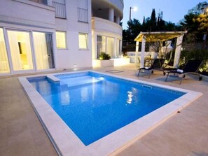 Une piscine rectangulaire bordée de carreaux, entourée d’une terrasse également carrelée, de couleur claire. On y trouve des chaises longues et une petite table avec un parasol.