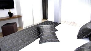 Ein modernes Hotelzimmer mit einem Bett, einem Flachbildfernseher, einem kleinen Tisch mit Flasche und Gläsern sowie einem Fenster mit Vorhängen.