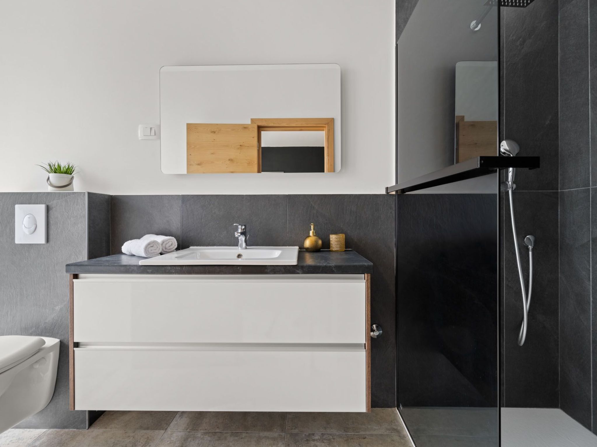 Een moderne badkamer met een douche in donkere tegels, een wit wastafelmeubel met een gouden kraan en een spiegel met houten lijst erboven.