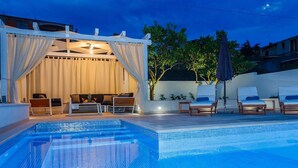 Una piscina con solárium empedrado, un área de estar techada con sofá y sillones, y vistas a árboles y edificios al fondo.