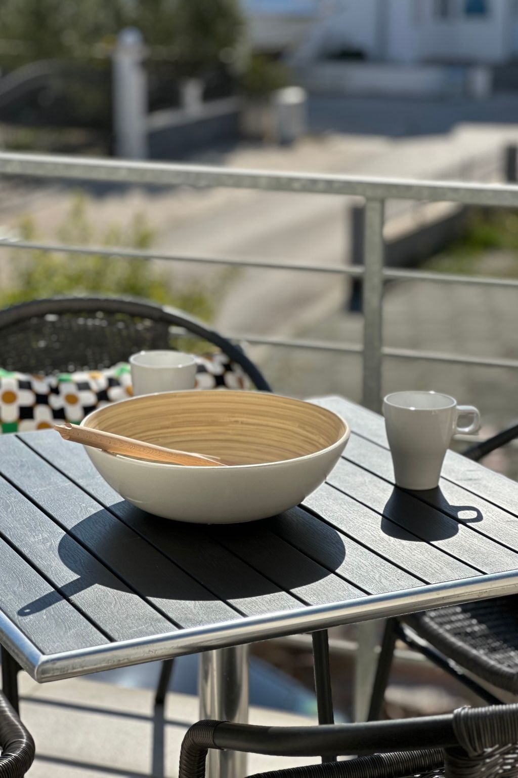 Una mesa con un tazón y una taza, una silla de mimbre y una barandilla al fondo.