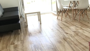 Een kamer met een houten vloer, een donkere bank, een eettafel met stoelen en een schuifdeur van glas die leidt naar een buitenruimte.