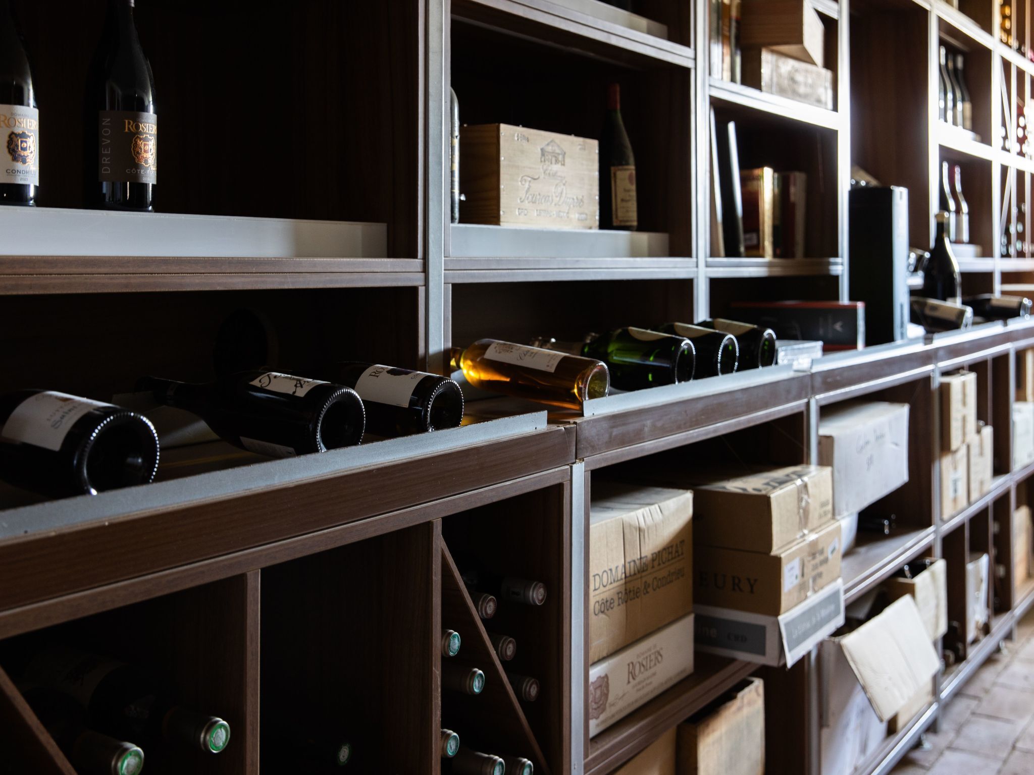 Une cave à vin avec des étagères remplies de bouteilles de vin et de cartons portant les noms des différents vins.