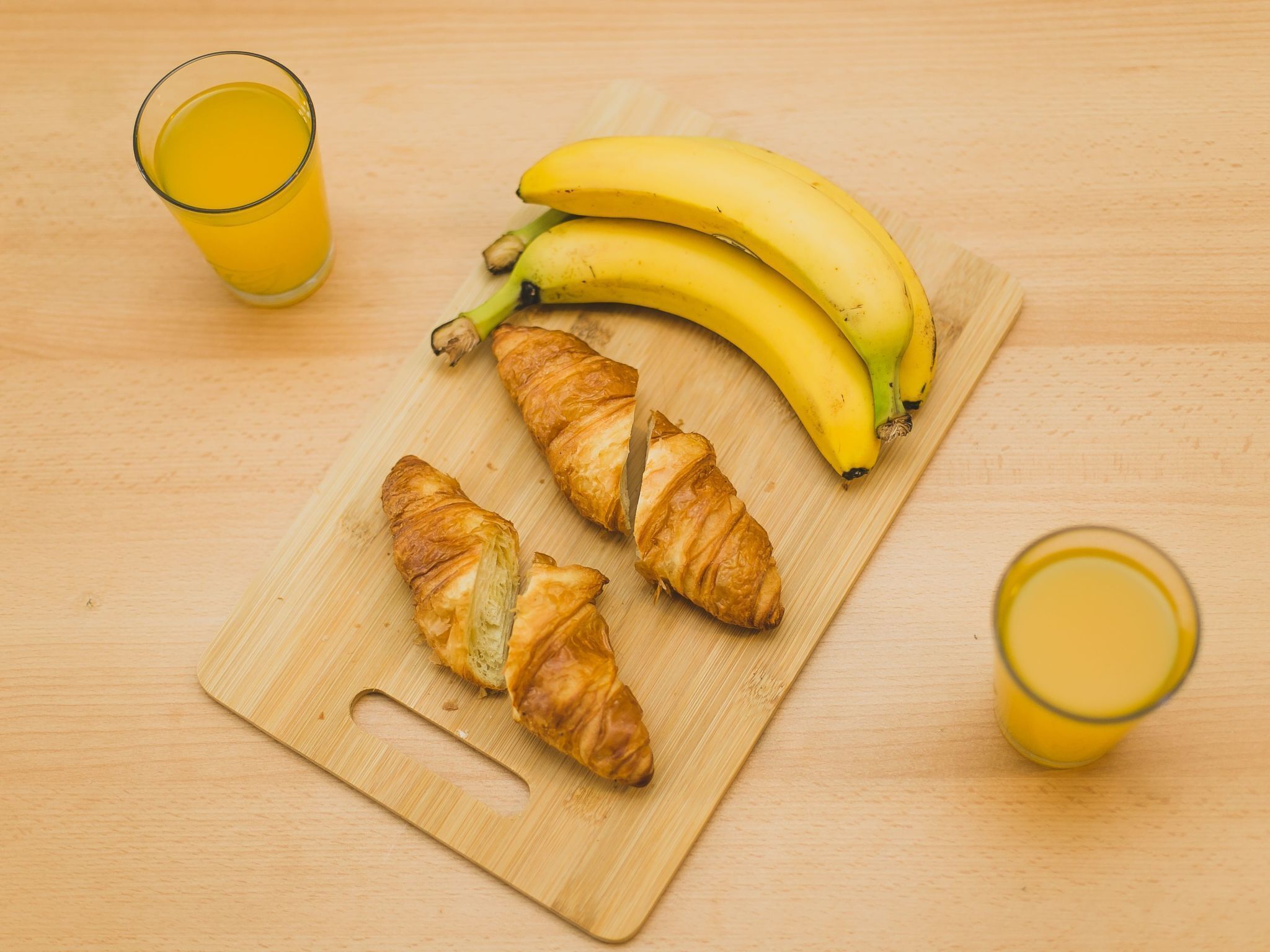 Una tabla de madera con dos croissants, un racimo de plátanos y dos vasos de jugo de naranja.