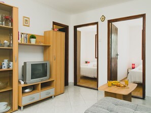 A compact living space with a television on a wooden stand, a bookshelf, a small table with a banana and a glass of orange juice, and a mirror reflecting a bed and a nightstand with a lamp. - Gelassenheit am Meer 1 by Interhome (Nin)