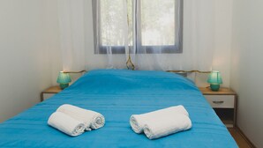 Un dormitori amb un llençol blau, dues tovalloles blanques, una finestra amb cortines de tul i dues làmpades de nit.