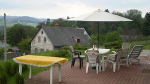 Une terrasse extérieure avec une table et des chaises, un grand parasol, et une maison en arrière-plan.