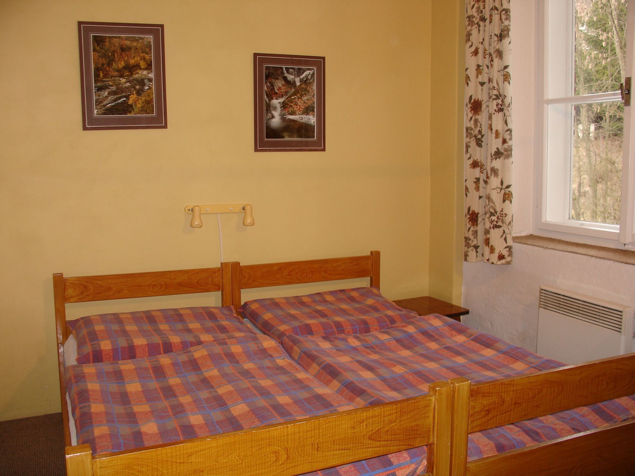 Een kamer met een houten bed, twee ingelijste foto's aan de muur, een raam met gordijnen en een radiator.