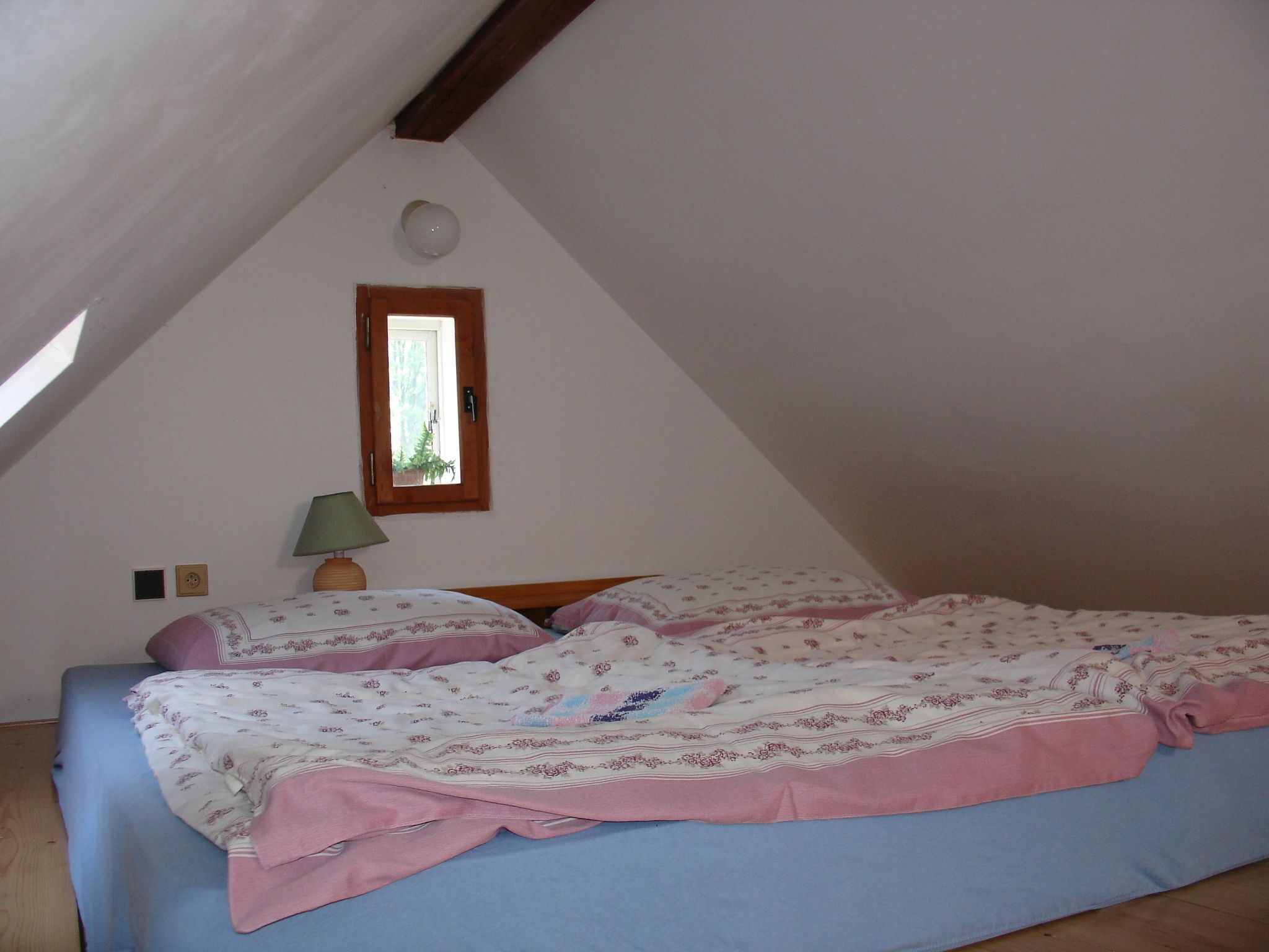 A bedroom with a sloped ceiling, a single bed with floral bedding, a small window with a view of greenery, and a bedside lamp.