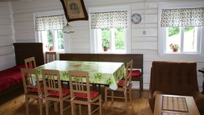 A dining area with a table set for six, surrounded by wooden chairs with red cushions. There are two windows with floral curtains, a wall clock, and a hanging light fixture. - Charmantes, traditionelles Holzhaus by Interhome (Jilemnice)