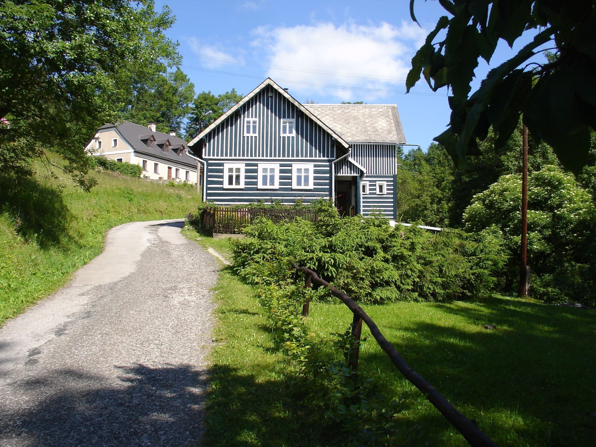 Une maison traditionnelle en bois, dotée d’un toit à pente, d’une allée en gravier et entourée d’une végétation luxuriante.