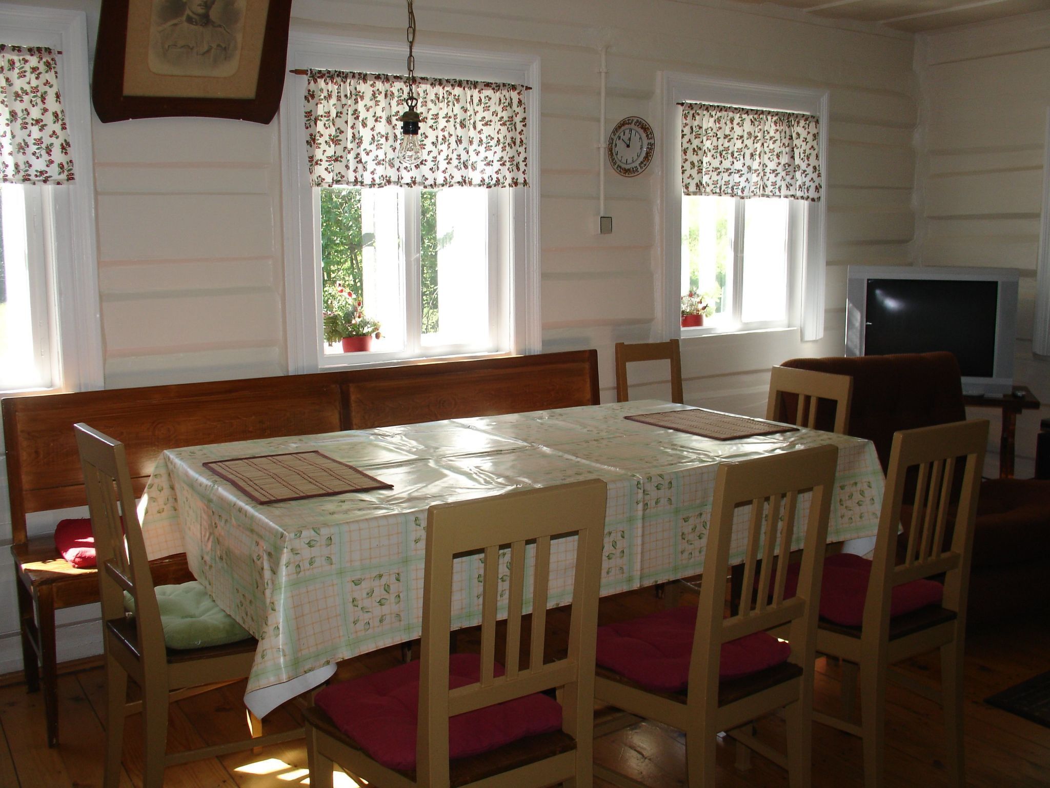 A dining room with a wooden table and chairs, a television, and windows with curtains.