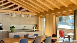 Eine moderne Küche mit Holzbalken, ein Essbereich mit großem Tisch und Stühlen und Blick auf eine verschneite Landschaft durch große Fenster.