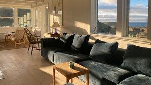 Ein gemütliches Wohnzimmer mit einem großen Fenster, das einen reizvollen Ausblick bietet, einem dunkelgrauen Sofa, Holzstühlen und einem kleinen Holztisch mit einer Lampe.