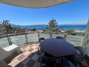 Terraço com piso xadrez, mesa e cadeiras, com vista para a costa, árvores e edifícios.