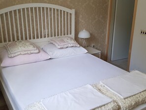 Een net opgemaakt bed met witte lakens en decoratieve kussens, een wit hoofdbord en een klein nachtkastje met een lamp.