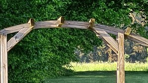 Puinen pergola, johon johtaa portaikko, sijaitsee hoidetun nurmikon ja tiheän lehtistön taustalla.