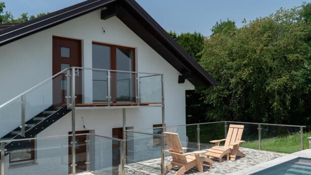 Ein modernes, zweistöckiges Haus mit einem Glasbalkon mit Blick auf einen Swimmingpool, ausgestattet mit Gartenmöbeln und einer Terrassenanlage mit Steinplatten.
