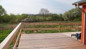 Un pontile in legno con ringhiera, un piccolo edificio con tetto arancione e vista su un paesaggio verde con alberi e arbusti.
