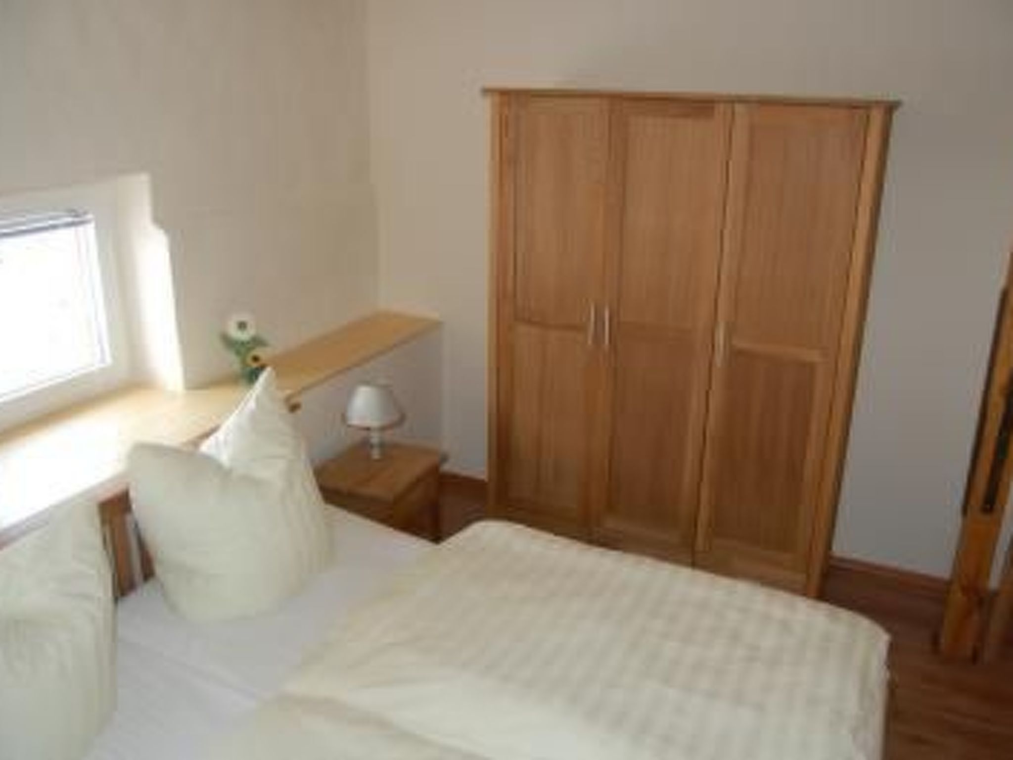 Une chambre à coucher avec un lit, des oreillers, une armoire en bois, une table de chevet avec une lampe et une fenêtre.
