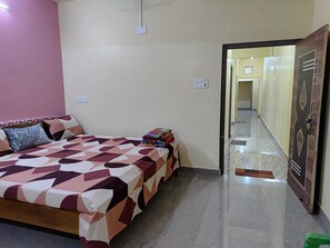 A bedroom with a bed, pillows, a bedside table with books, and a door leading to a hallway. - Laxmi Nrusingha Bhawan (Puri)
