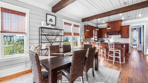 Un'ampia sala da pranzo con un tavolo in legno scuro e sedie in vimini, una cucina con mobili in legno e illuminazione a sospensione, e ampie finestre con tende.