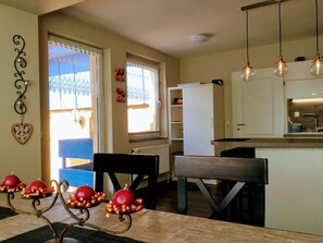 Un comedor con una mesa dispuesta para cuatro, una zona de cocina con refrigerador, y un ventanal con cortinas decorativas.