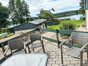 Un patio con sillas de mimbre y una mesa de vidrio, con vista a un lago que tiene un muelle y un edificio verde al fondo.