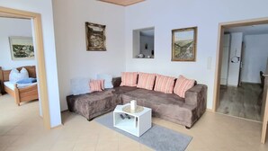 Ein Wohnzimmer mit einem braunen Ecksofa, einem weißen Couchtisch und einem grauen Teppich. An der Wand hängen drei gerahmte Bilder.