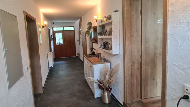 En gang med en trædør for enden, et lille bord med en vase med tørrede planter, og en reol med bøger og pynteting.