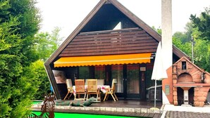 A wooden cabin with a sloped roof, a covered patio with outdoor seating, and a small brick structure to the right. - in Kuhklingen mit Großer Terrasse by Interhome (Wald-Michelbach)