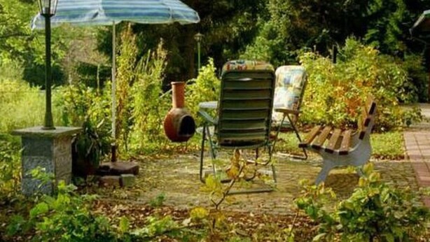 Un patio agrémenté d’un jardin, avec un parasol bleu, un lampadaire en pierre, une chaise rustique et un banc en bois.