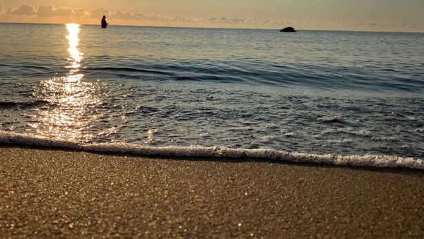 Ein Strand mit ruhiger See, einer sichtbaren Welle und der untergehenden Sonne im Hintergrund.