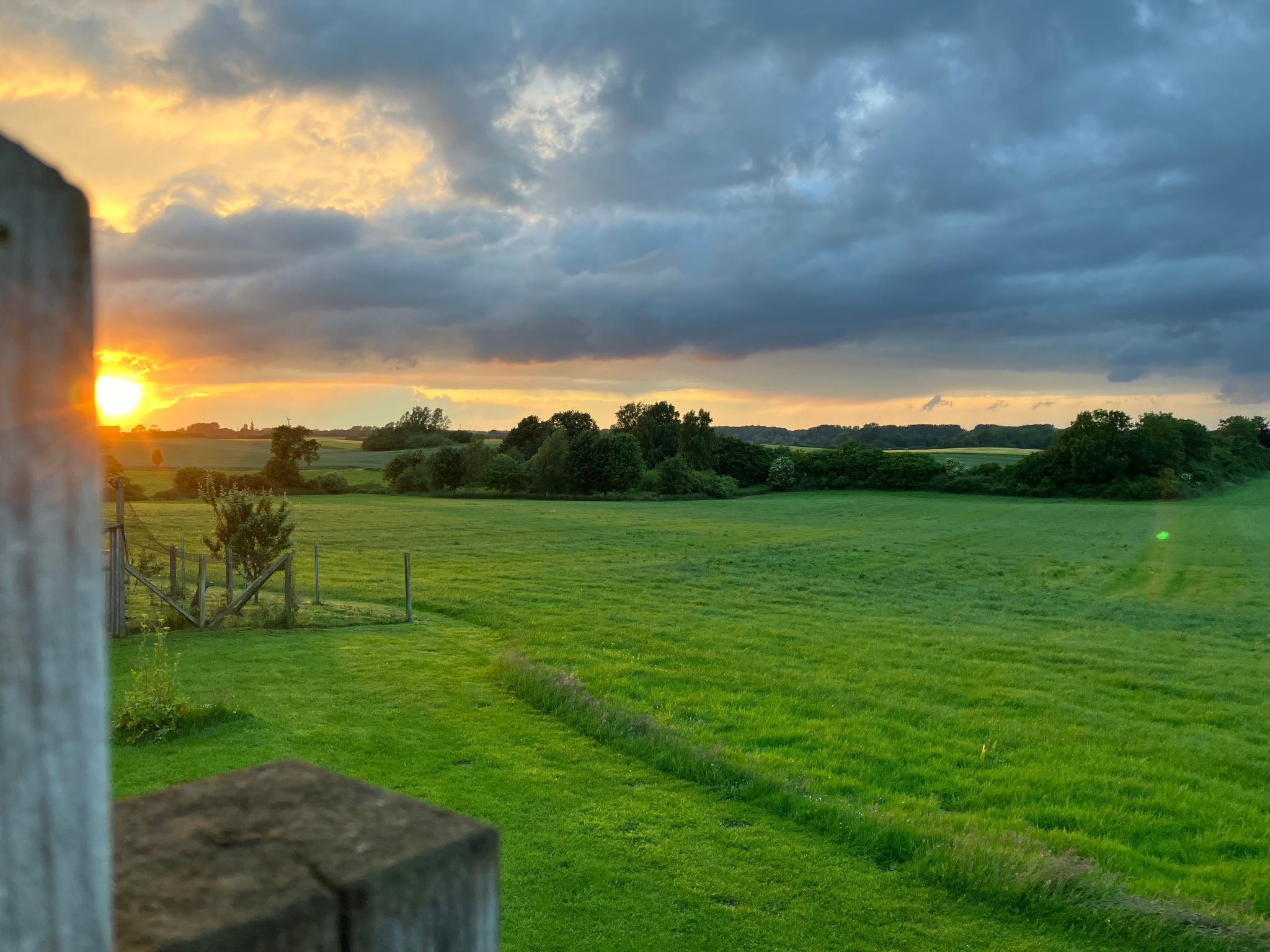A rural landscape with a field and a fence in the foreground, and a cloudy sky with the sun setting in the background.