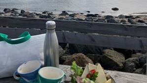Une table en bois, avec un thermos, deux tasses et un bol de nourriture, placée près d’une plage rocheuse, avec vue sur la mer.