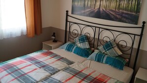 Un letto rifatto con una coperta a quadri e cuscini abbinati, una lampada da comodino, un comodino e un quadro raffigurante una foresta sulla parete.