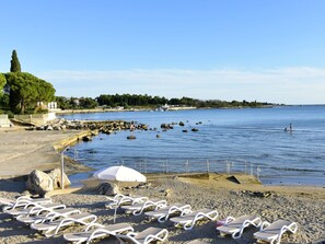 A beach with white lounge chairs and a white umbrella, rocky shoreline, and calm water. - Vesna Peovic by Interhome (Umag/Zambratija)