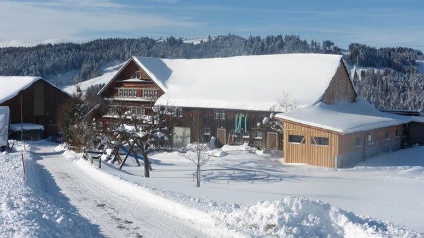 Un chalet en bois, dont le toit est recouvert de neige, avec un télésiège à proximité et une forêt en arrière-plan.