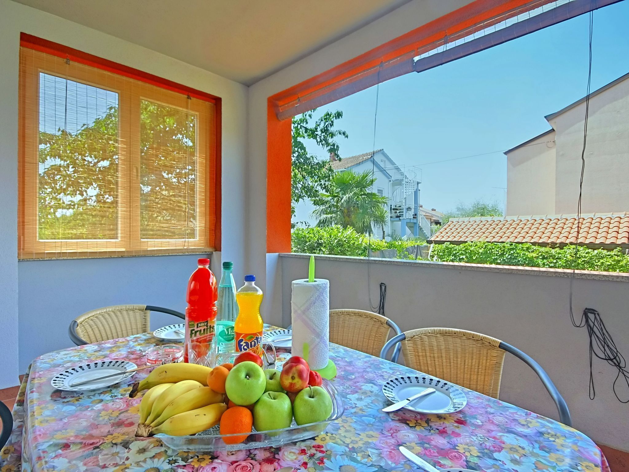 Une table de balcon ornée d’une nappe à motifs floraux, avec des fruits, des boissons, et une vue sur une maison dont le toit est recouvert de tuiles.