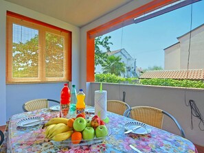 Une table de balcon ornée d’une nappe à motifs floraux, avec des fruits, des boissons, et une vue sur une maison dont le toit est recouvert de tuiles.