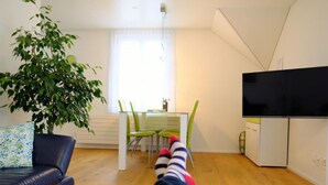 Ein Wohnzimmer mit einer Couch, einem Esstisch mit Stühlen, einem Fernseher und einer großen Pflanze.