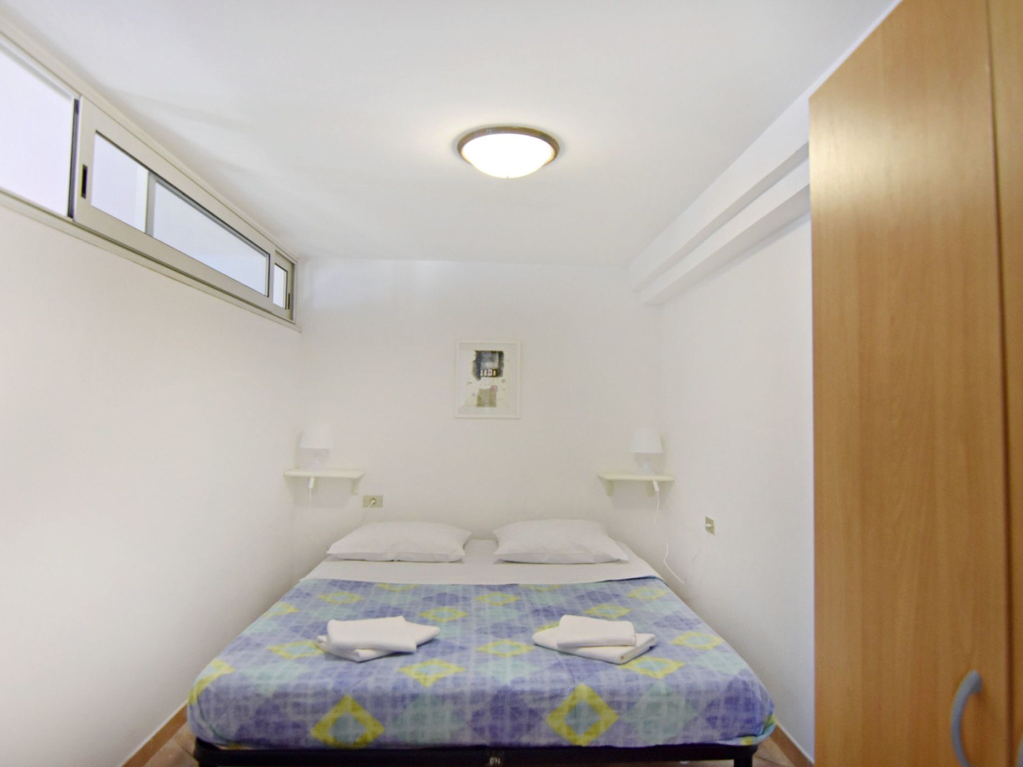 Ein kleines Zimmer mit einem Einzelbett, einem Fenster, einem Nachttisch und einem Holzschrank.